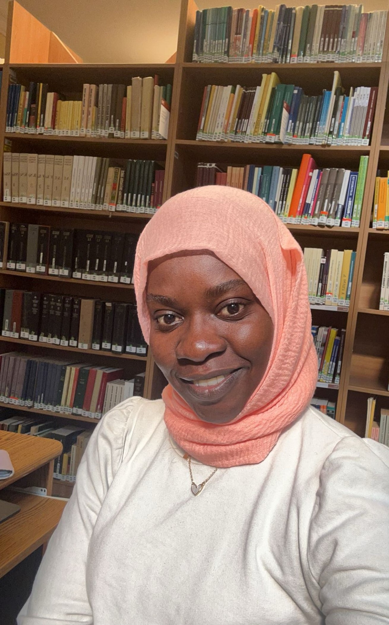 Photo of Nafisa wearing a pink headscarf and white top, smiling at the camera, with many shelves of books in the background.