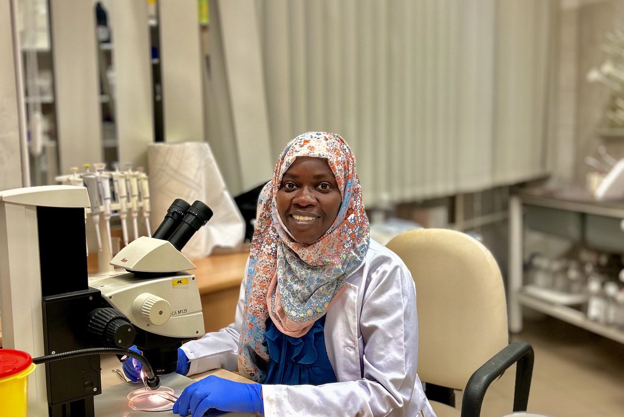 Nafisa wearing a flowery-patterned headscarf, a white lab coat and blue gloves, sitting at a desk with a microscope on it