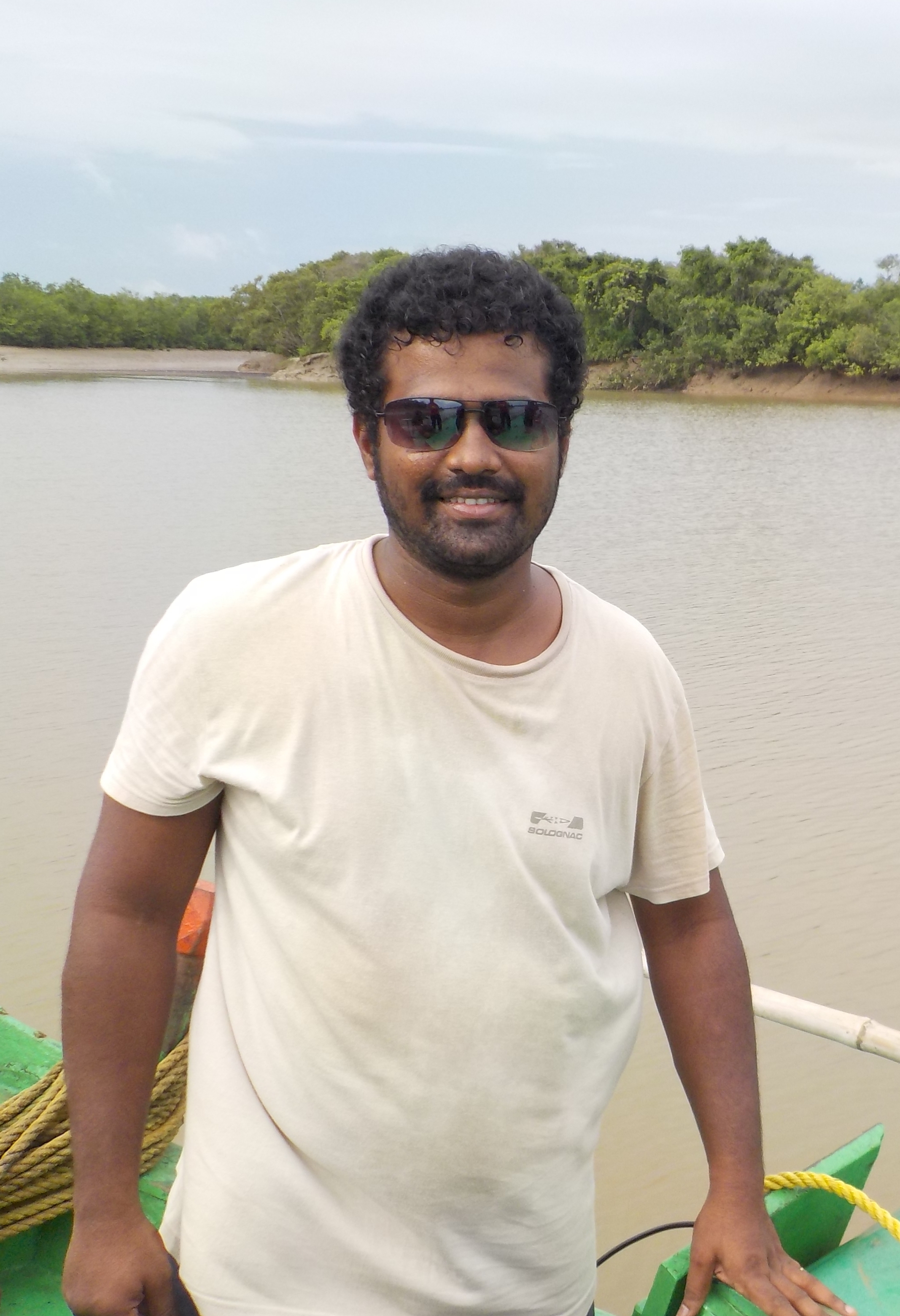 Ravi wearing sunglasses smiling at the camera. He is standing, with a body of water and mud banks and vegetation in the background.