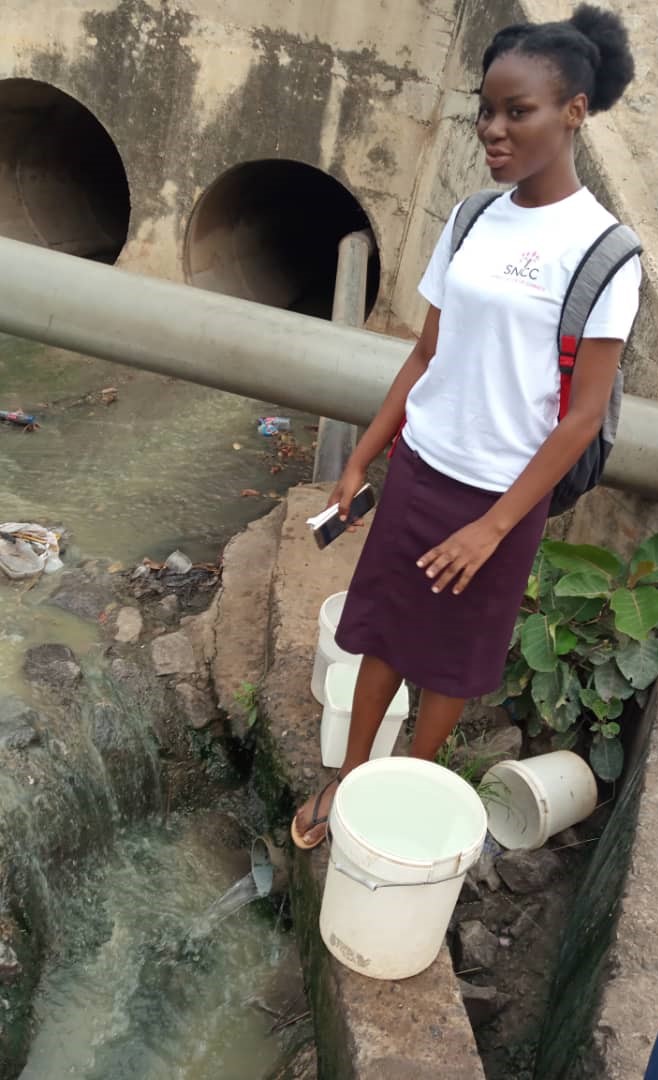 Bless stands on a low wall at the side of running water flowing from two very large pipe shaped structures. White buckets are around her feet. Bless wears a white t-shit and purple skirt.